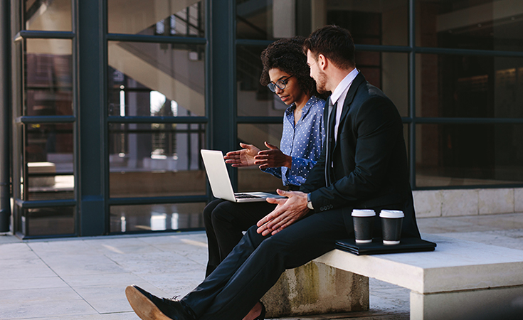 Two business people sitting on a bench in modern office building with a laptop. Woman with laptop discussing business with male colleague.
