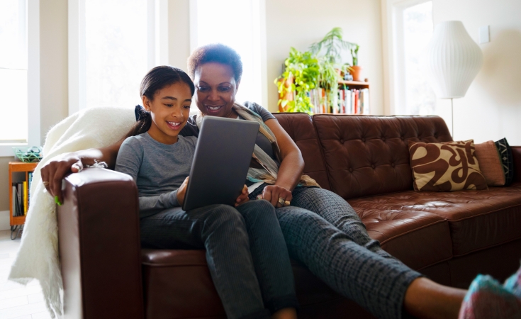 Mother and daughter smiling while looking at a tablet and sitting on a sofa