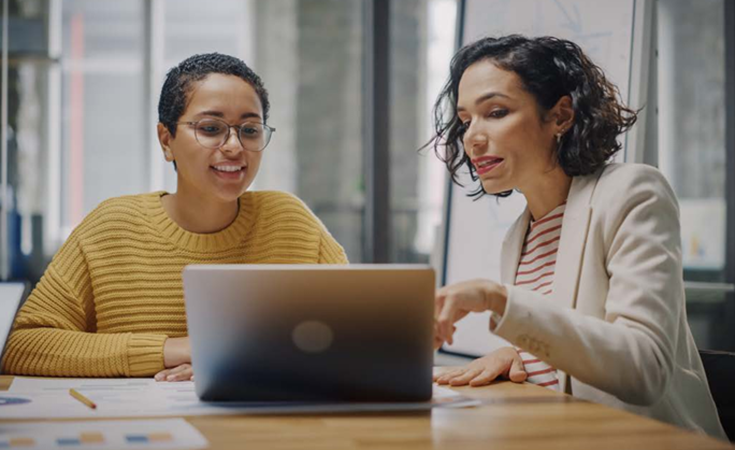 two women siting at a conference table pointing and looking at a computer screen