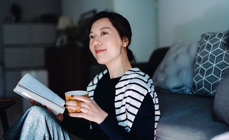 Beautiful smiling young Asian woman relaxing at home, reading a book with a cup of tea. Having a technology-free moment. Enjoying a quiet time and relaxing moment at cozy home. Healthy lifestyle living
